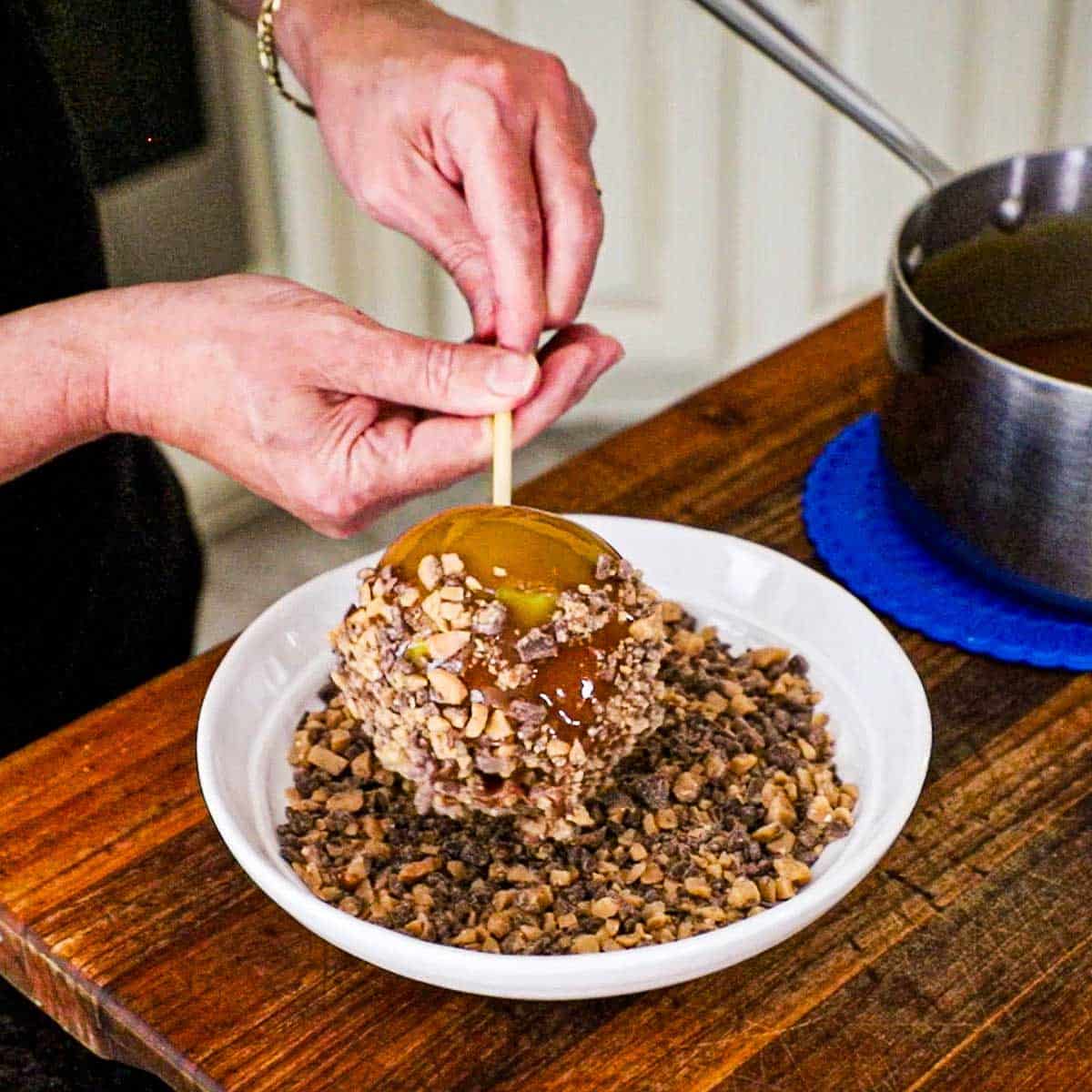 A person holding an inserted wooden stick into a honey caramel apple and is rolling the sticky apple through a shallow bowl filled with toffee bits.