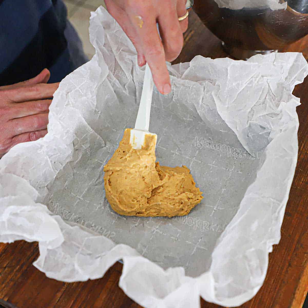 A person using a rubber spatula to smear pumpkin spice coffee cake batter into a square pan that is lined with white parchment paper.