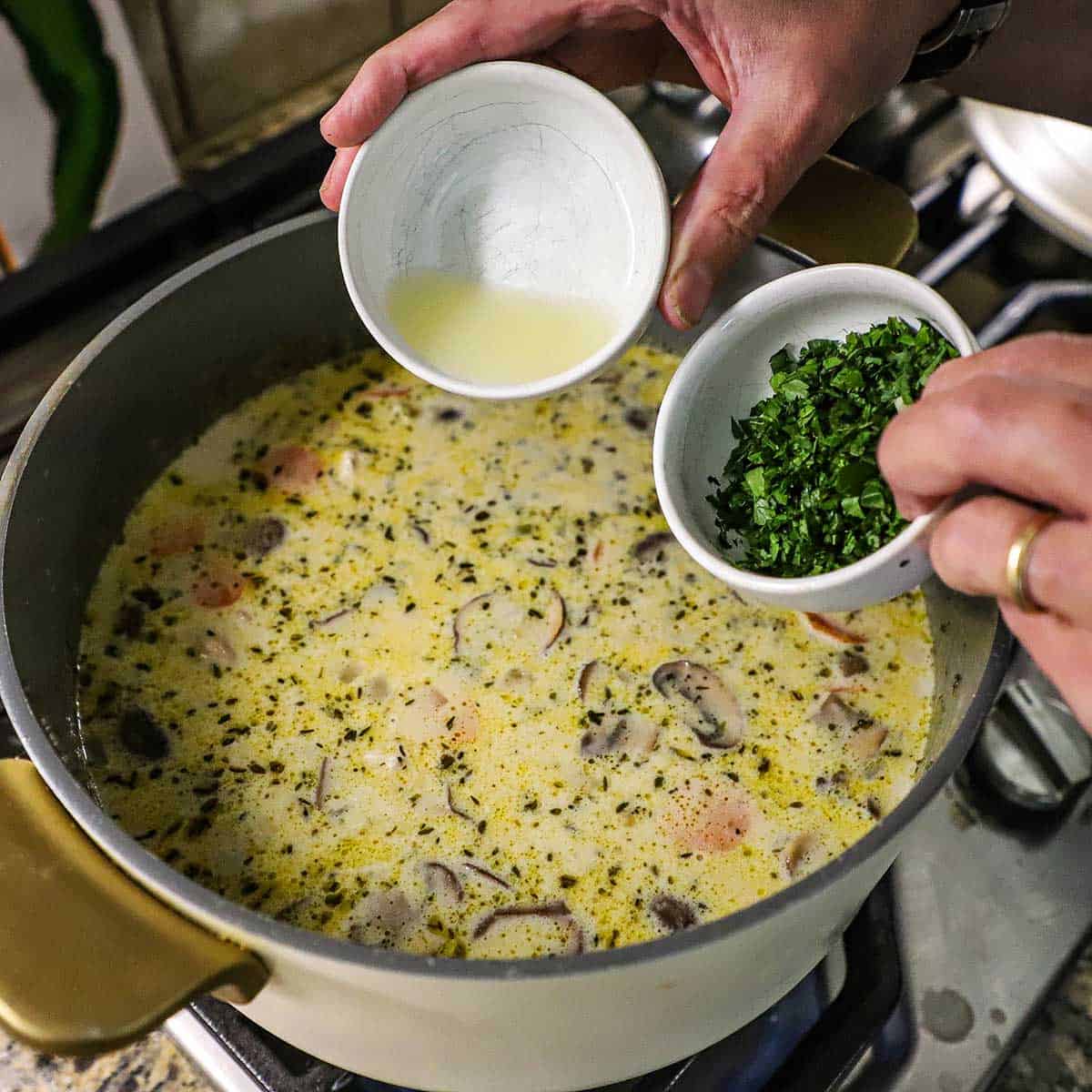 A person holding a small white bowl filled with fresh lemon juice in one hand and another small white bowl filled with chopped parsley in the other hand both over a pot of simmering creamy chicken and wild rice soup on a gas stove.