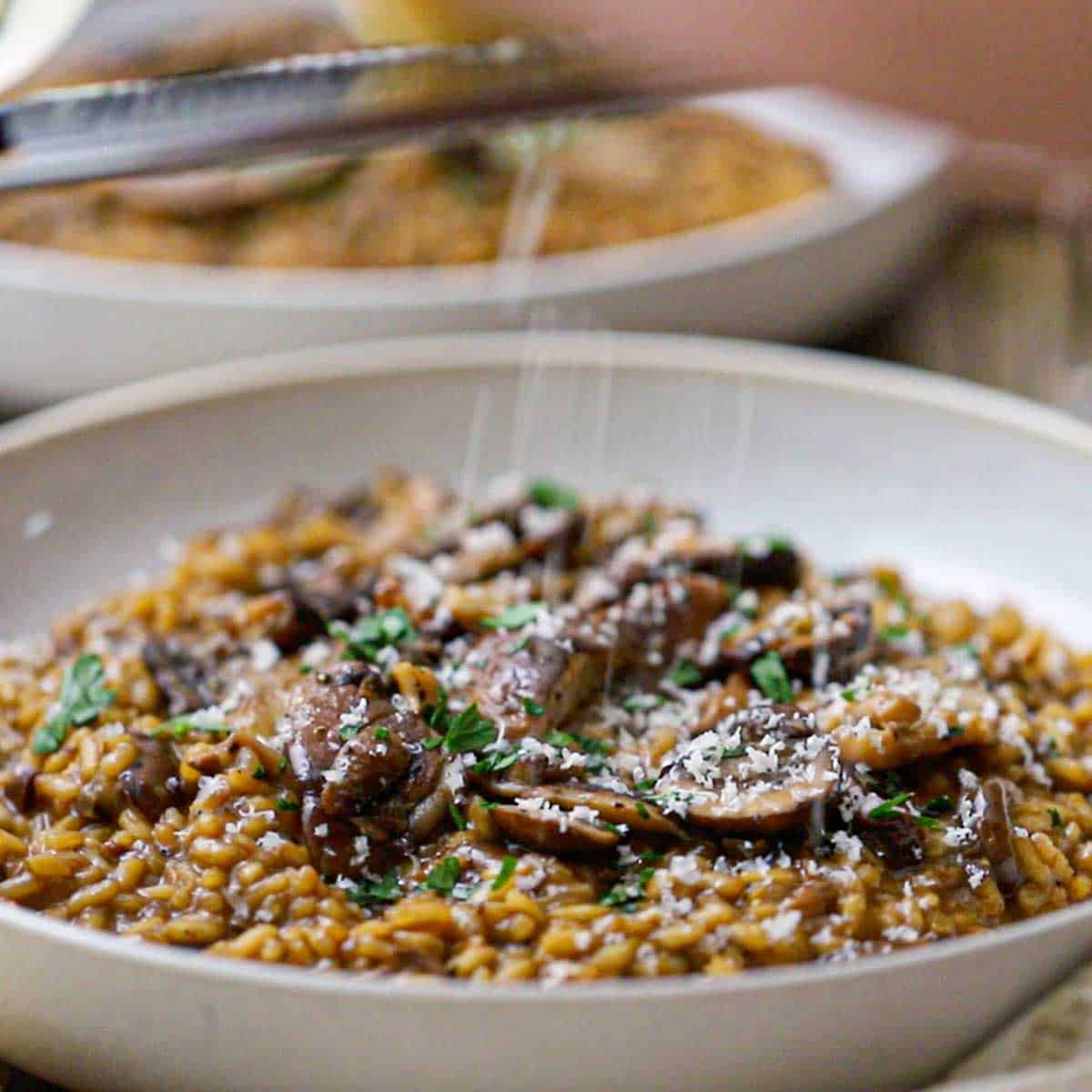 A person using a microplane to grate Parmesan cheese over the top of a shallow pasta bowl filled with a serving of wild mushroom risotto.