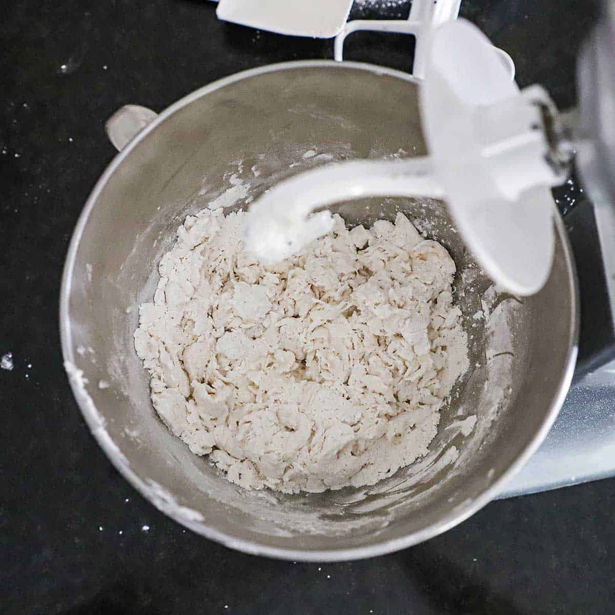 An overhead view of a shaggy dough that has just formed in the bowl of a stand mixer with the dough hook attached.