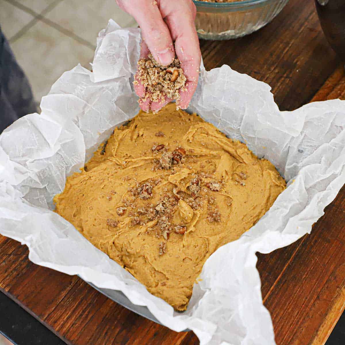 A person using his hand to sprinkle a pecan streusel over a layer pumpkin spice coffee cake batter that is has been spread over parchment paper that is lining a square baking pan.