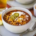 A straight-on view of a white soup bowl filled with a serving of Weeknight Beef Chili garnished with shredded cheddar cheese, chopped onions, sour cream, and sliced pickled jalapeño peppers.