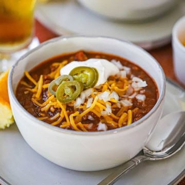 A straight-on view of a white soup bowl filled with a serving of Weeknight Beef Chili garnished with shredded cheddar cheese, chopped onions, sour cream, and sliced pickled jalapeño peppers.