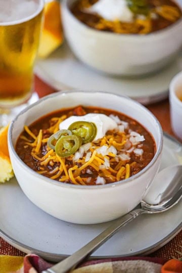 A straight-on view of a white soup bowl filled with a serving of Weeknight Beef Chili garnished with shredded cheddar cheese, chopped onions, sour cream, and sliced pickled jalapeño peppers.