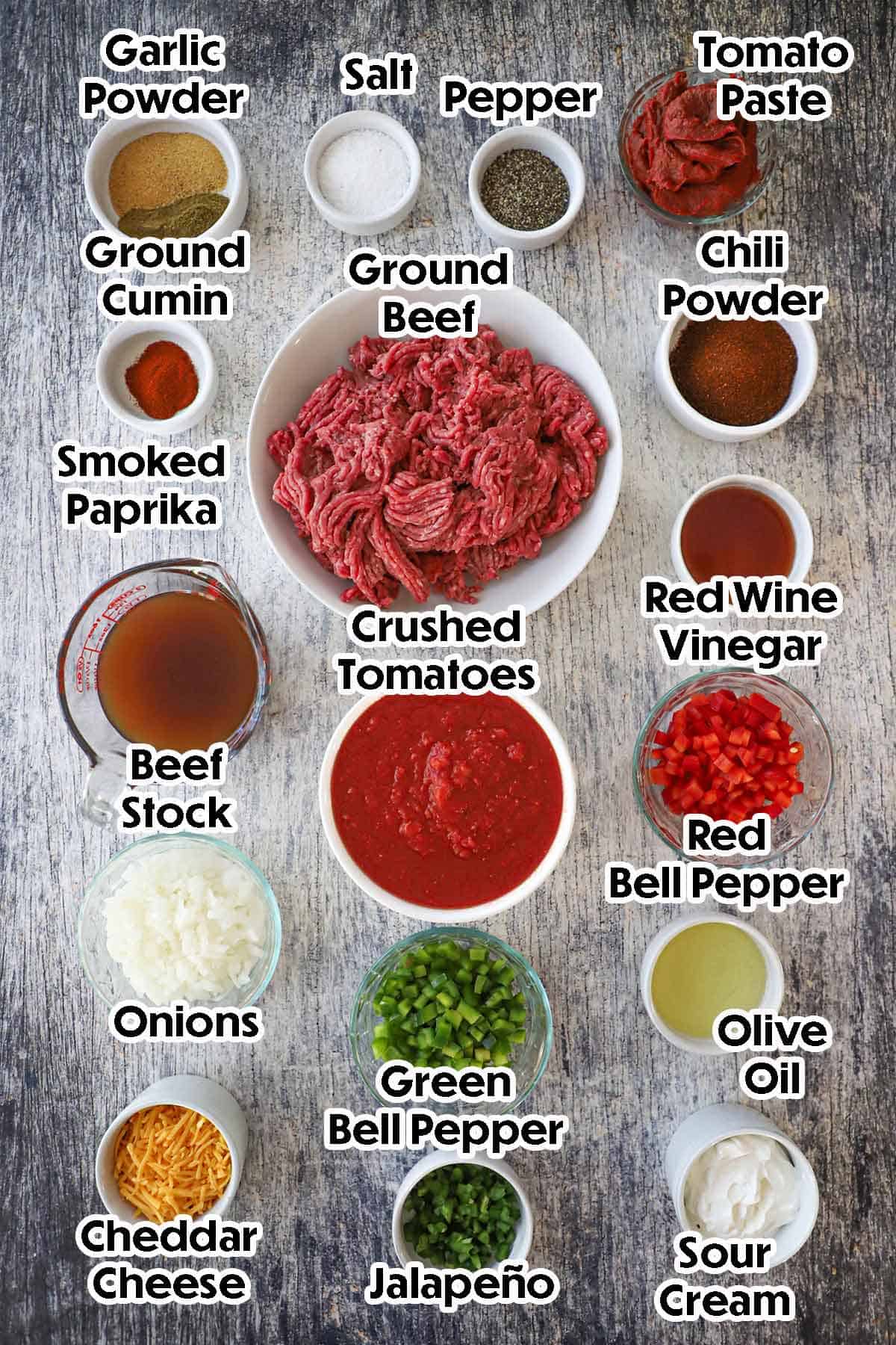 An arrangement of ingredients for Weeknight Beef Chili on a grey wooden background including ground beef, crushed tomatoes, chopped onions and peppers, beef stock, vinegar, tomato paste, oil, and seasonings.