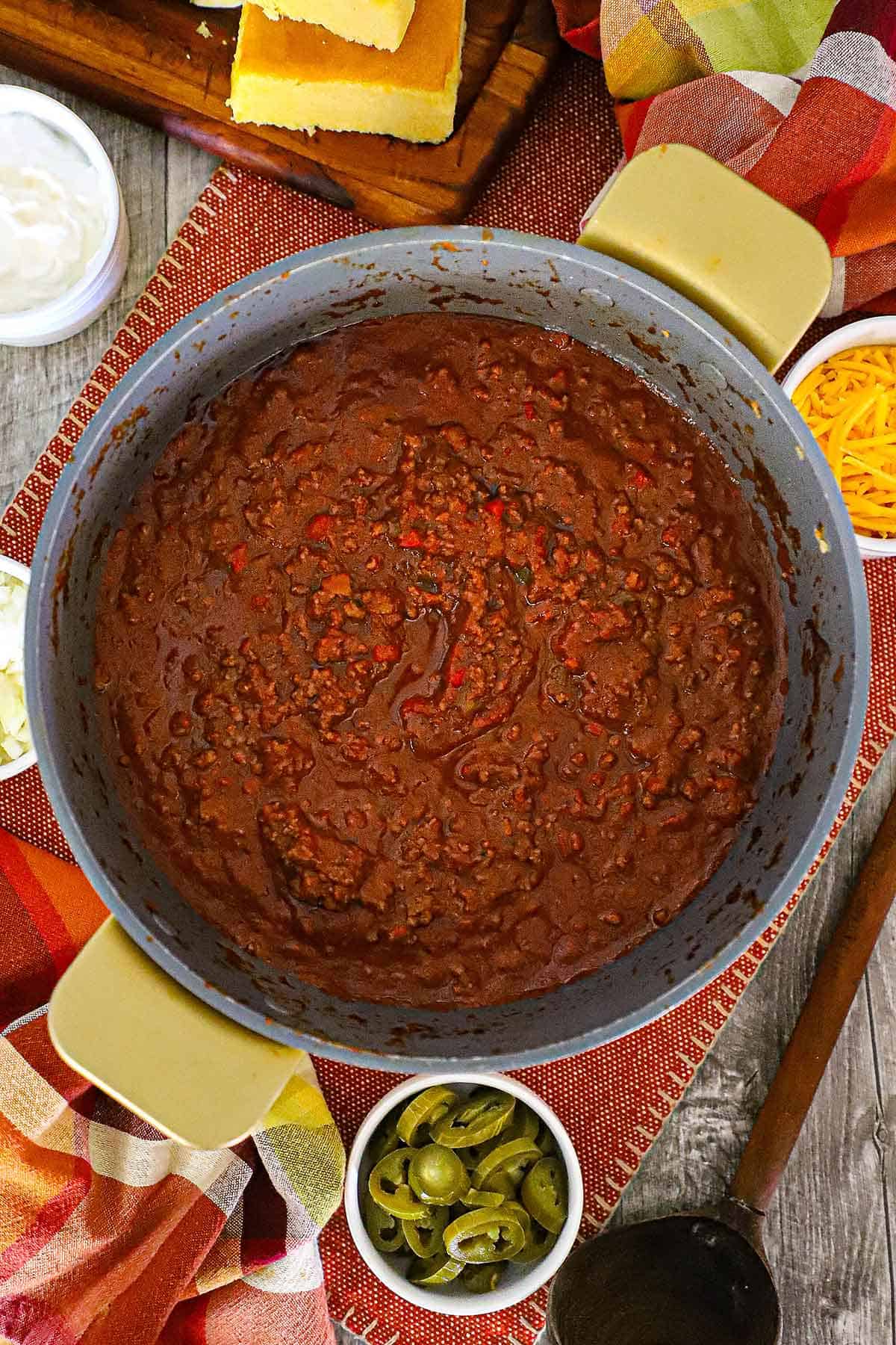 An overhead view of a large pot filled with a freshly prepared batch of Weeknight Beef Chili with a small bowl of sliced jalapeños and cornbread both nearby.