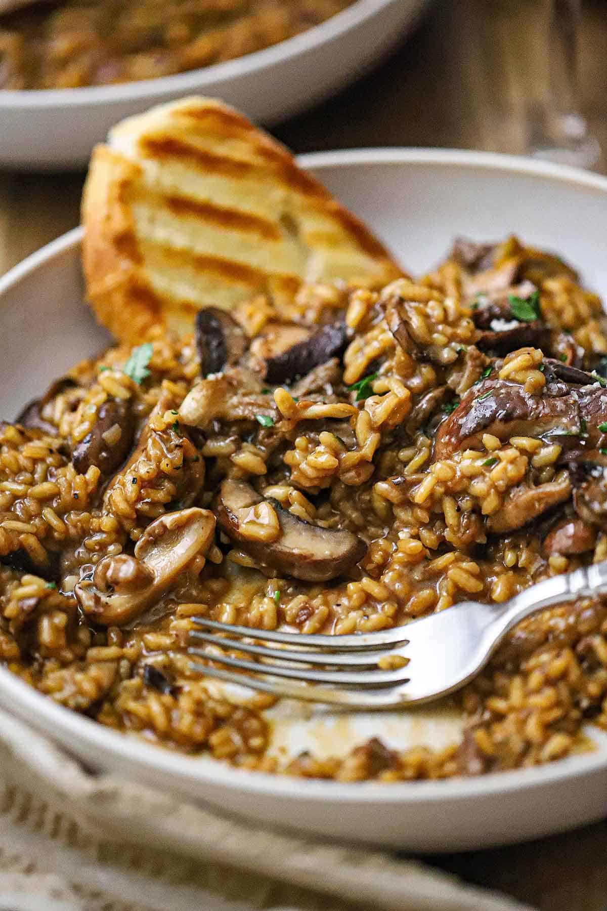 A straight-on view of a shallow pasta bowl filled with a partially eaten serving of wild mushroom risotto with a grilled slice of bread resting in the back of the bowl.