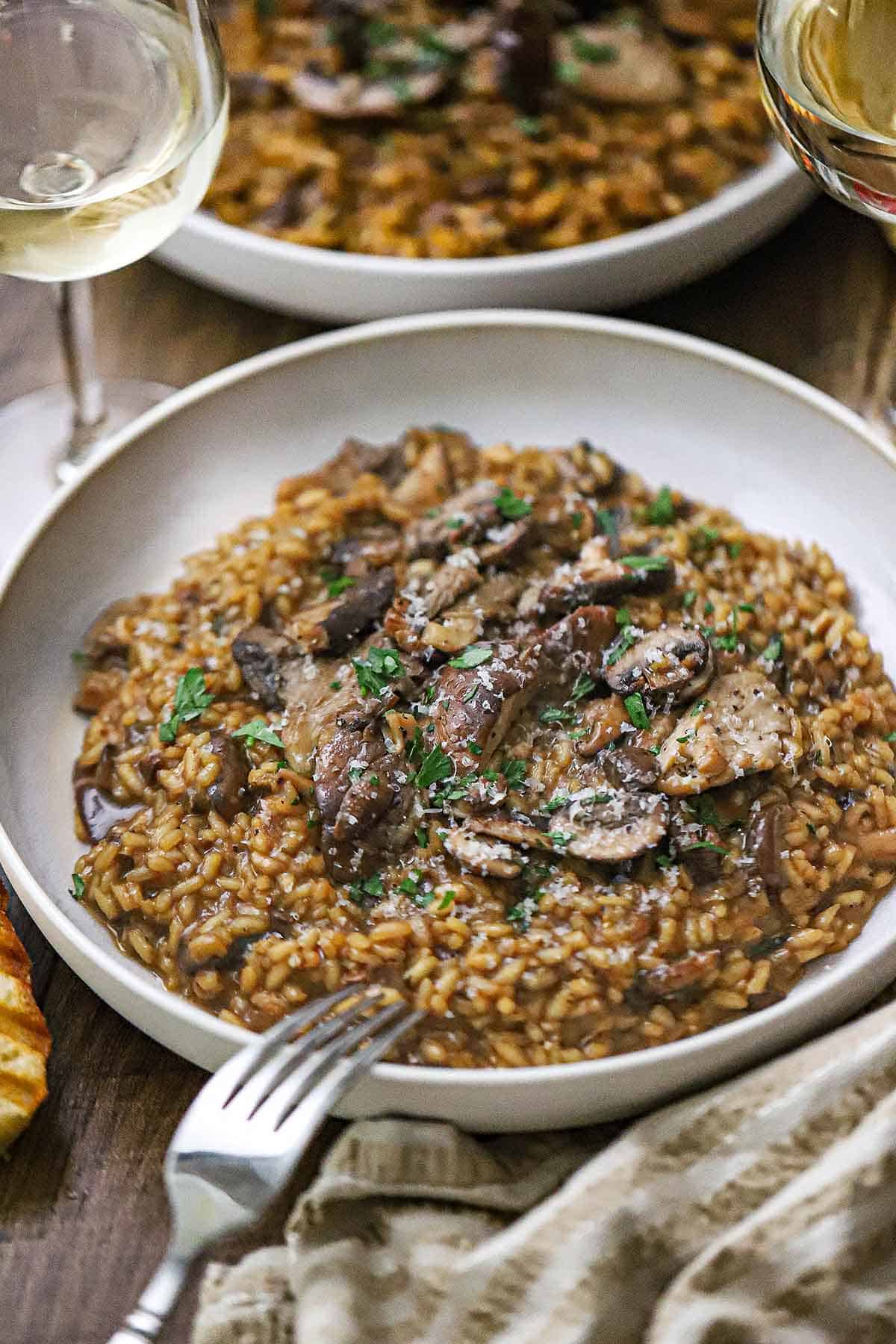 A straight-on view of two shallow pasta bowls each filled with a serving of wild mushroom risotto garnished with grated Parmesan cheese and chopped parsley.